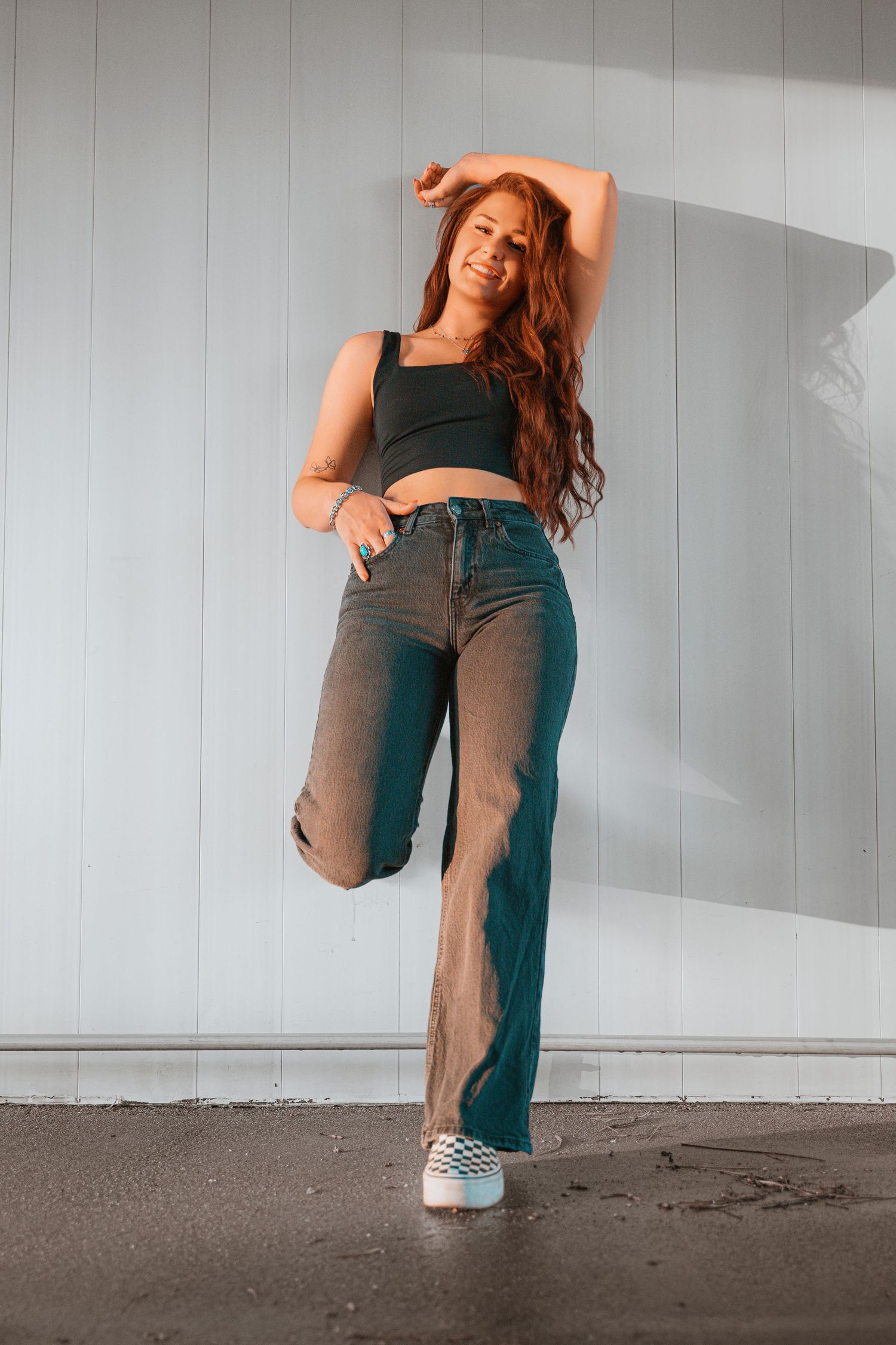 Young woman with long red hair poses in crop top and wide-leg jeans against white wall.