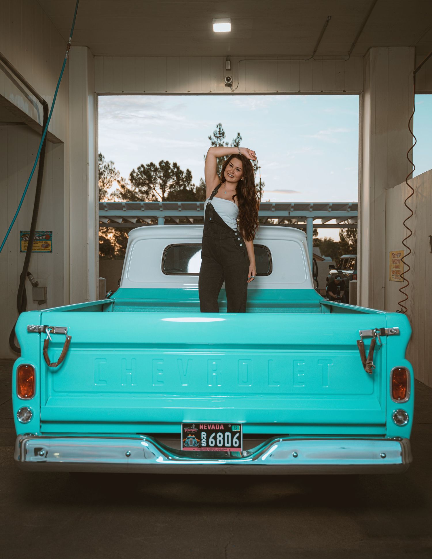 Woman posing on the bed of a classic teal Chevrolet pickup truck in a garage at sunset.