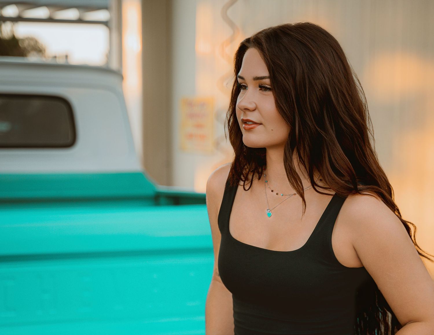 Young woman with long dark hair wearing a black tank top and turquoise necklace standing outdoors near a teal wall.