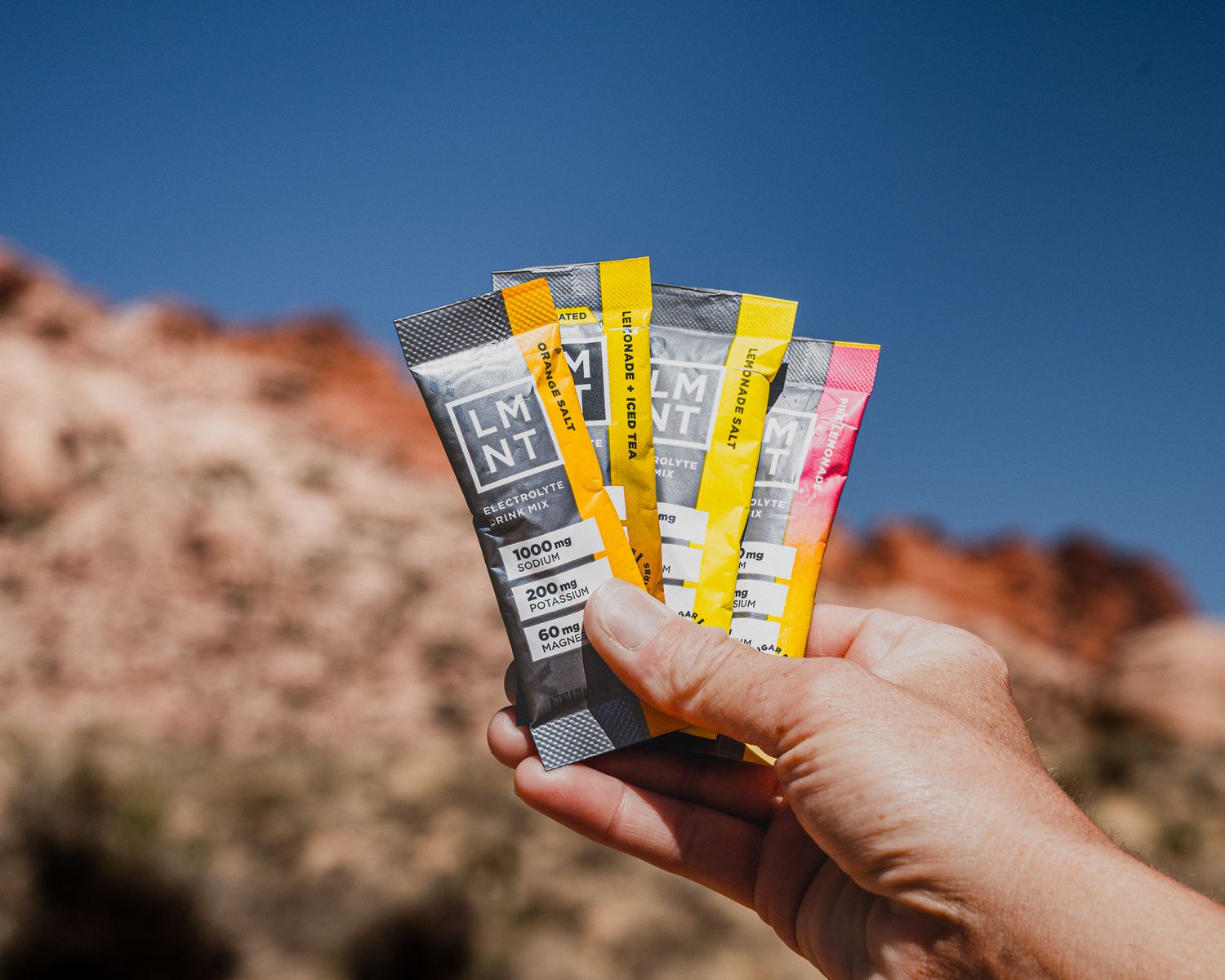 Hand holding four LMNT electrolyte drink mix packets in assorted flavors against a blue desert sky.