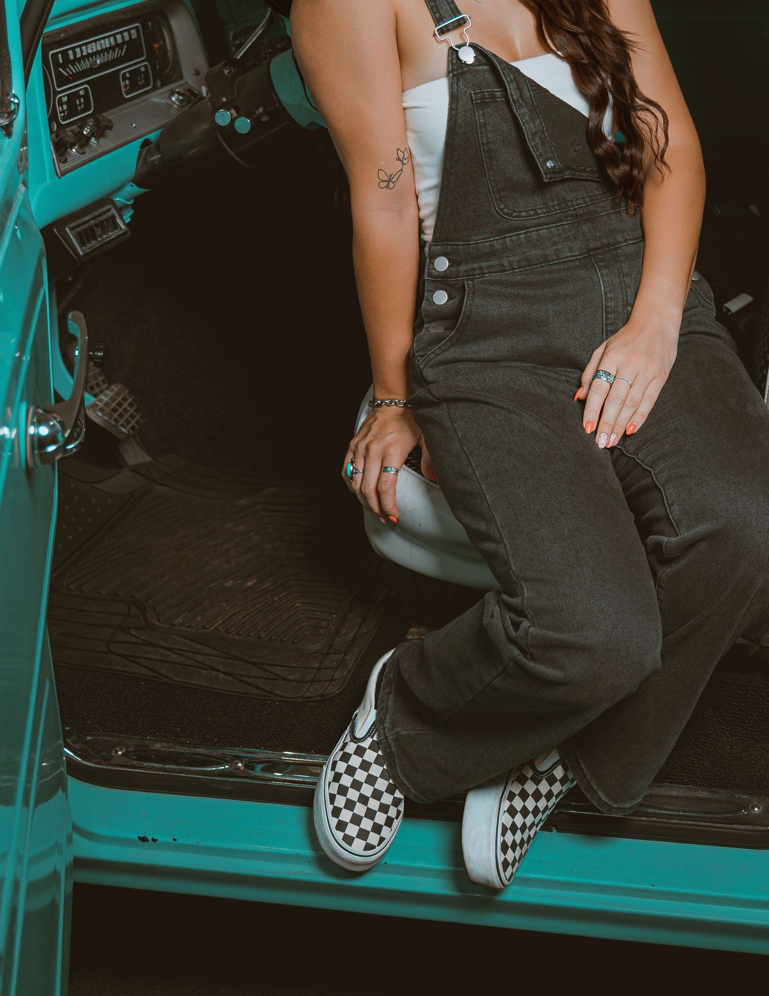 Woman in black denim overalls and checkered Vans slip-ons sitting in a teal vintage vehicle.