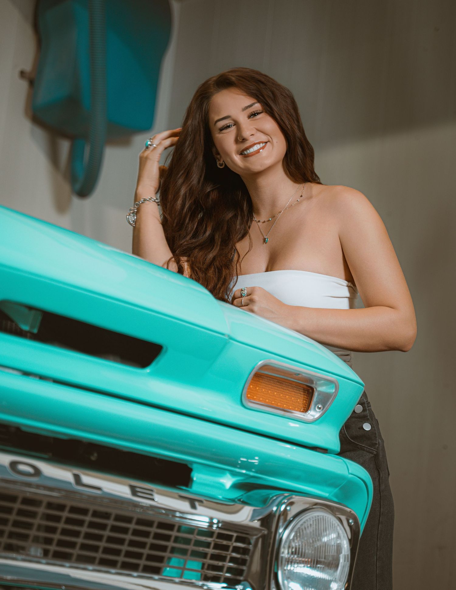 Smiling brunette woman in white tube top leaning on a vintage turquoise Chevrolet truck hood.