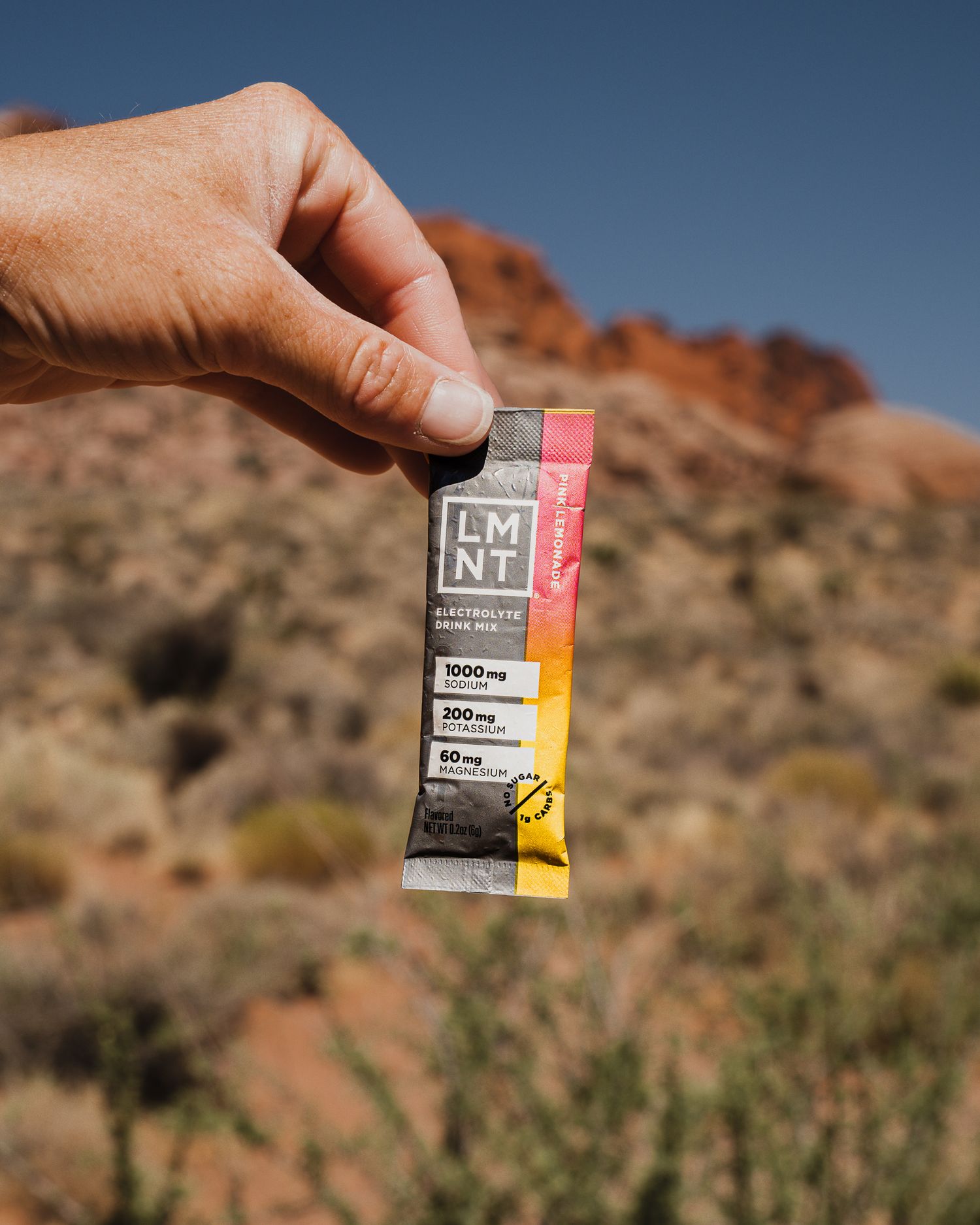 Hand holding an LMNT electrolyte drink mix packet against a bright blue sky and red rock desert backdrop.