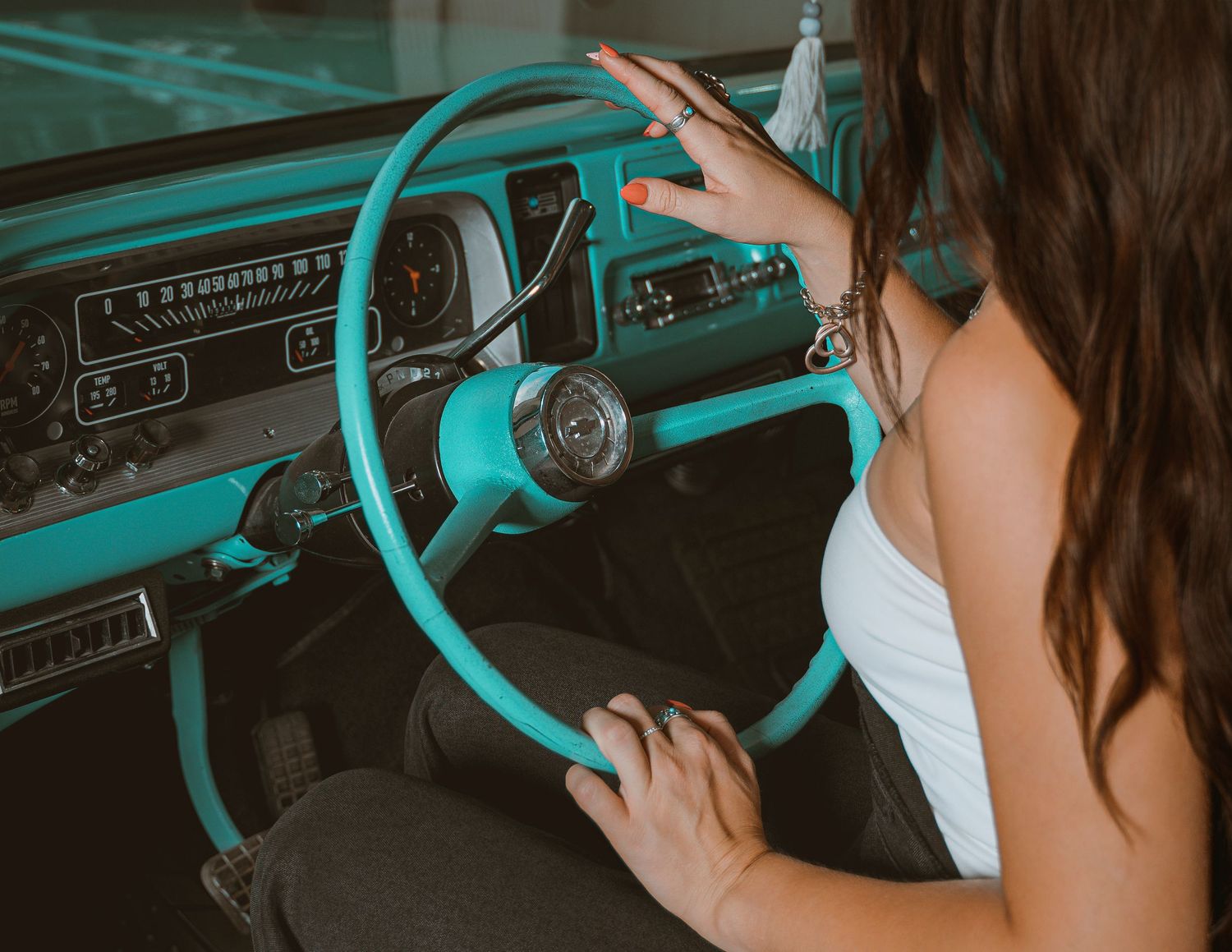 Woman with long brown hair sitting in a vintage teal car, hands on the turquoise steering wheel.