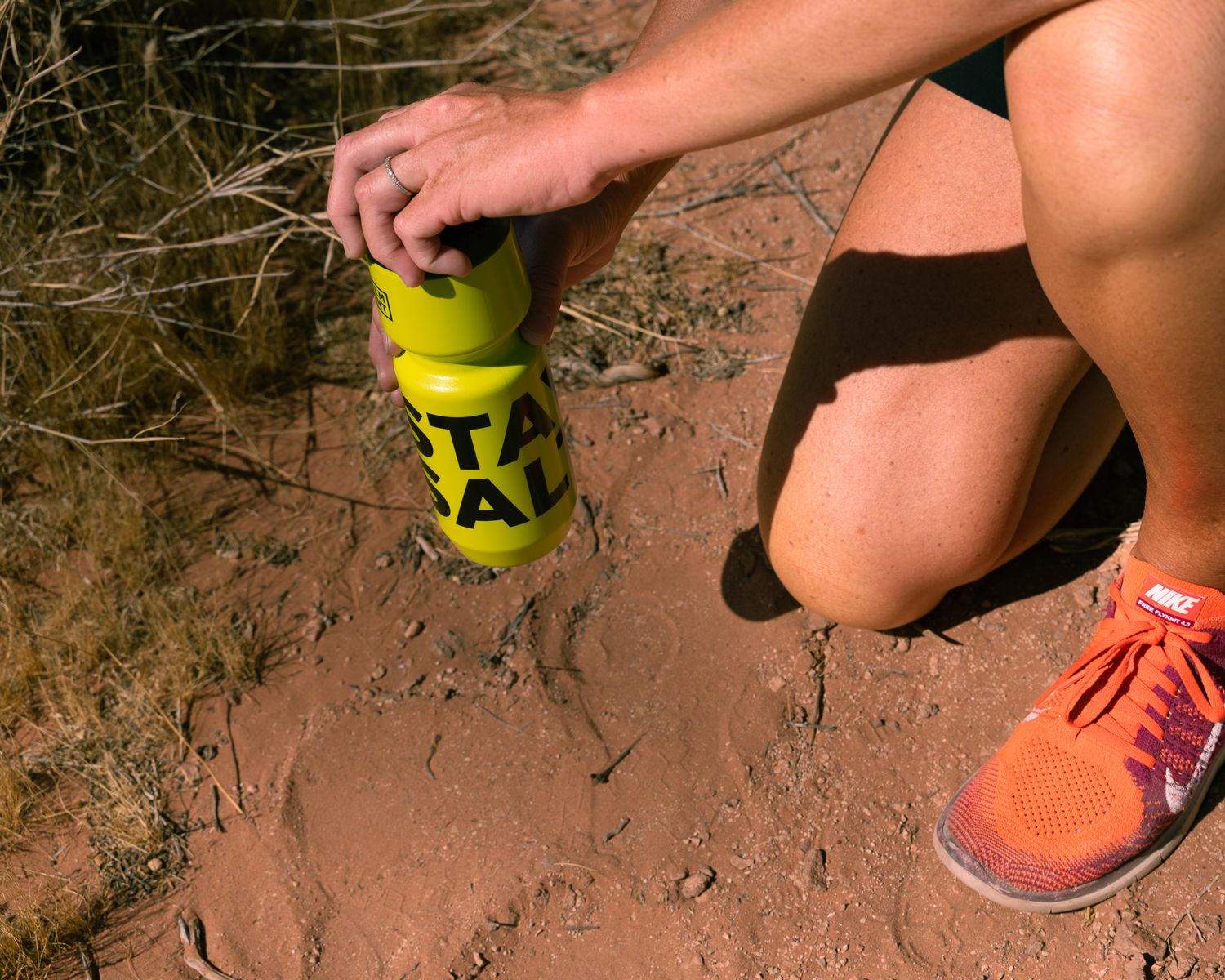 Close-up of hands opening a yellow STAY SALTY LMNT bottle on a red dirt trail with orange running shoes.