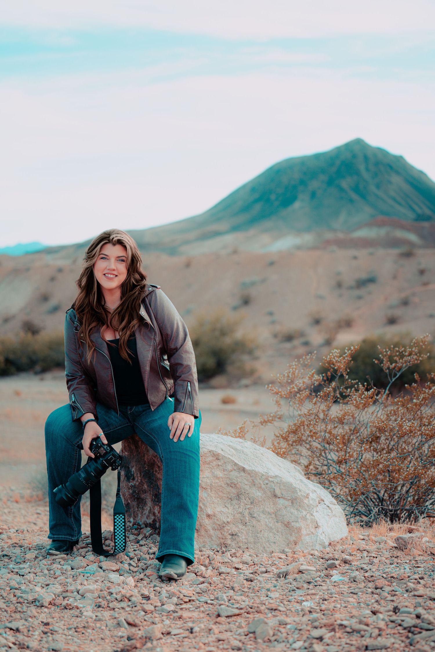 Woman photographer sitting on a rock in a desert landscape, holding a camera, with a volcanic mountain in the background.