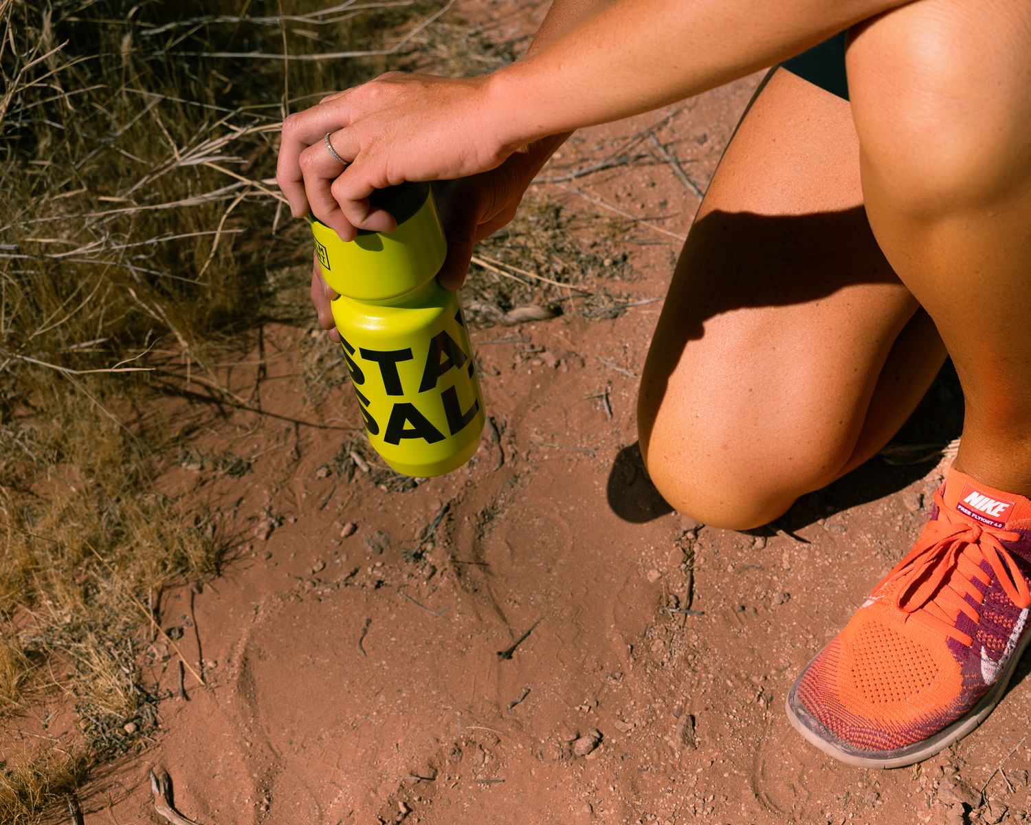 Female trail runner in orange sneakers opening a yellow STAY SALTY water bottle on red dirt ground.