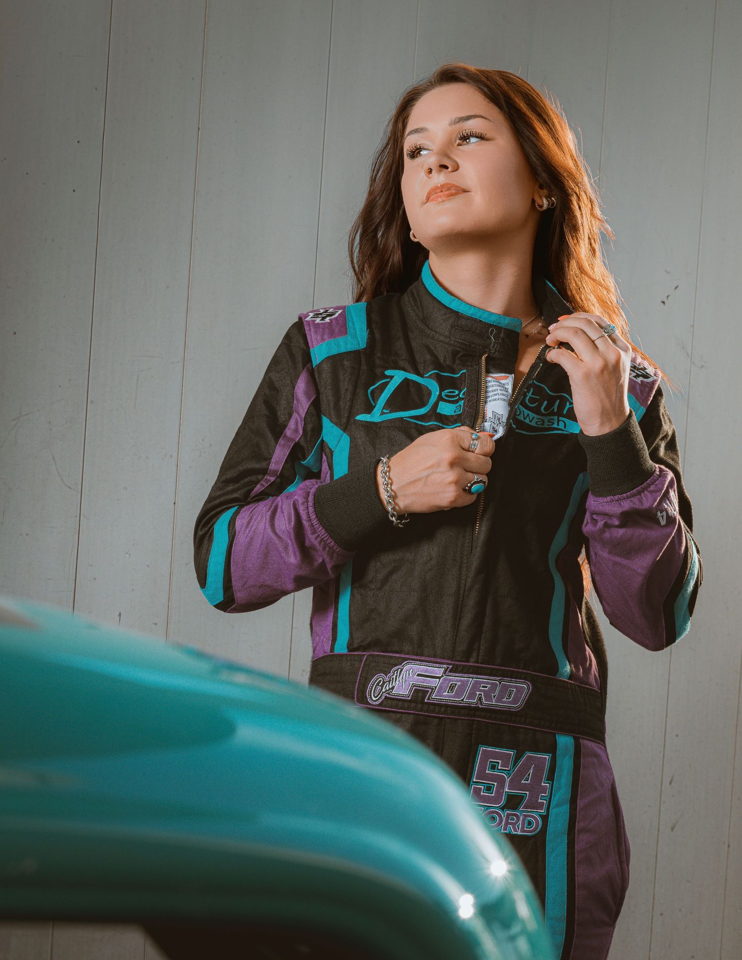 Young woman in black racing suit standing beside teal vintage car outdoors.