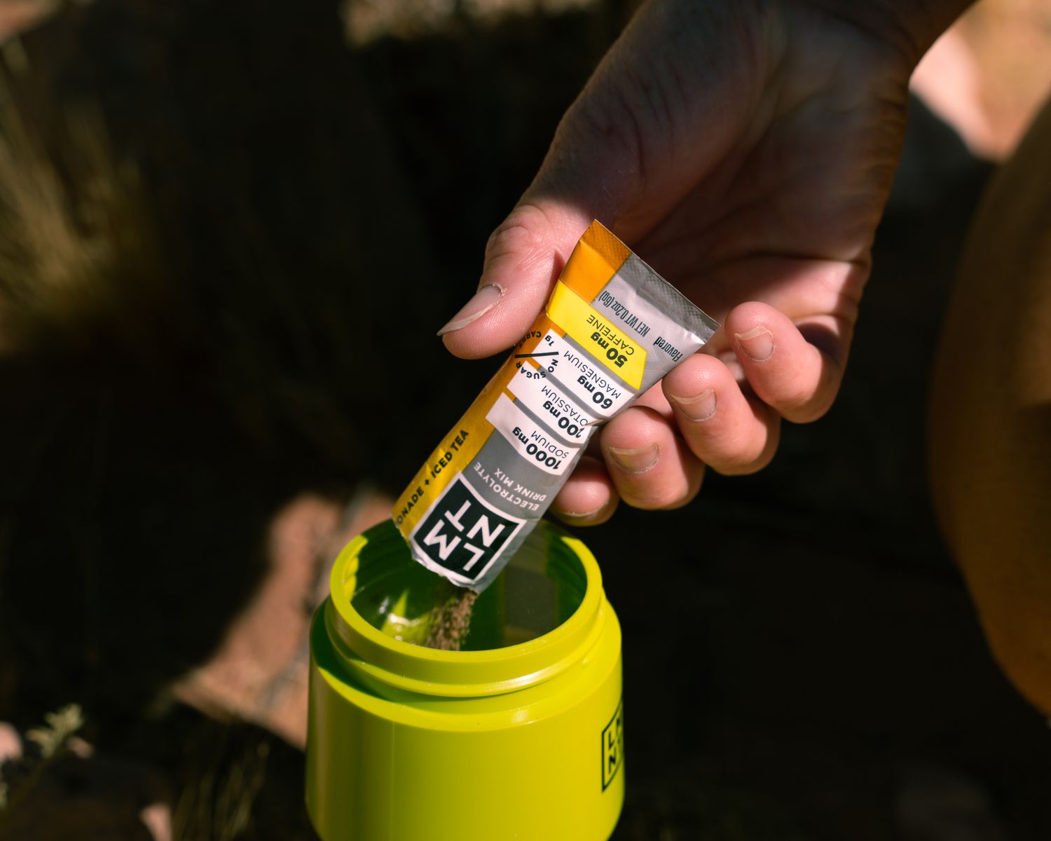 Hand pouring LMNT electrolyte powder packet into a yellow STAY SALTY water bottle outdoors.