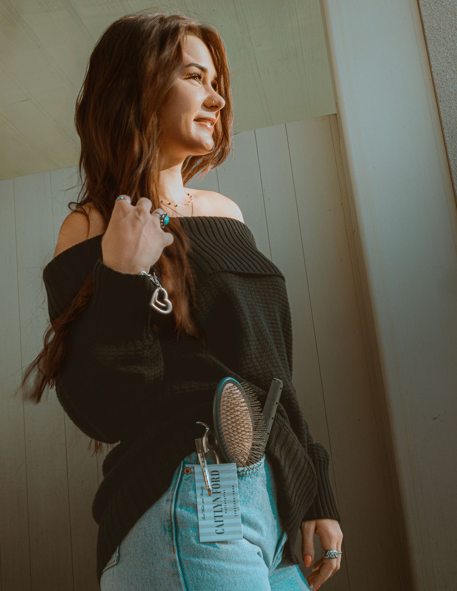 Young woman in black off-shoulder knit sweater with turquoise jewelry and jeans posing in sunlight.