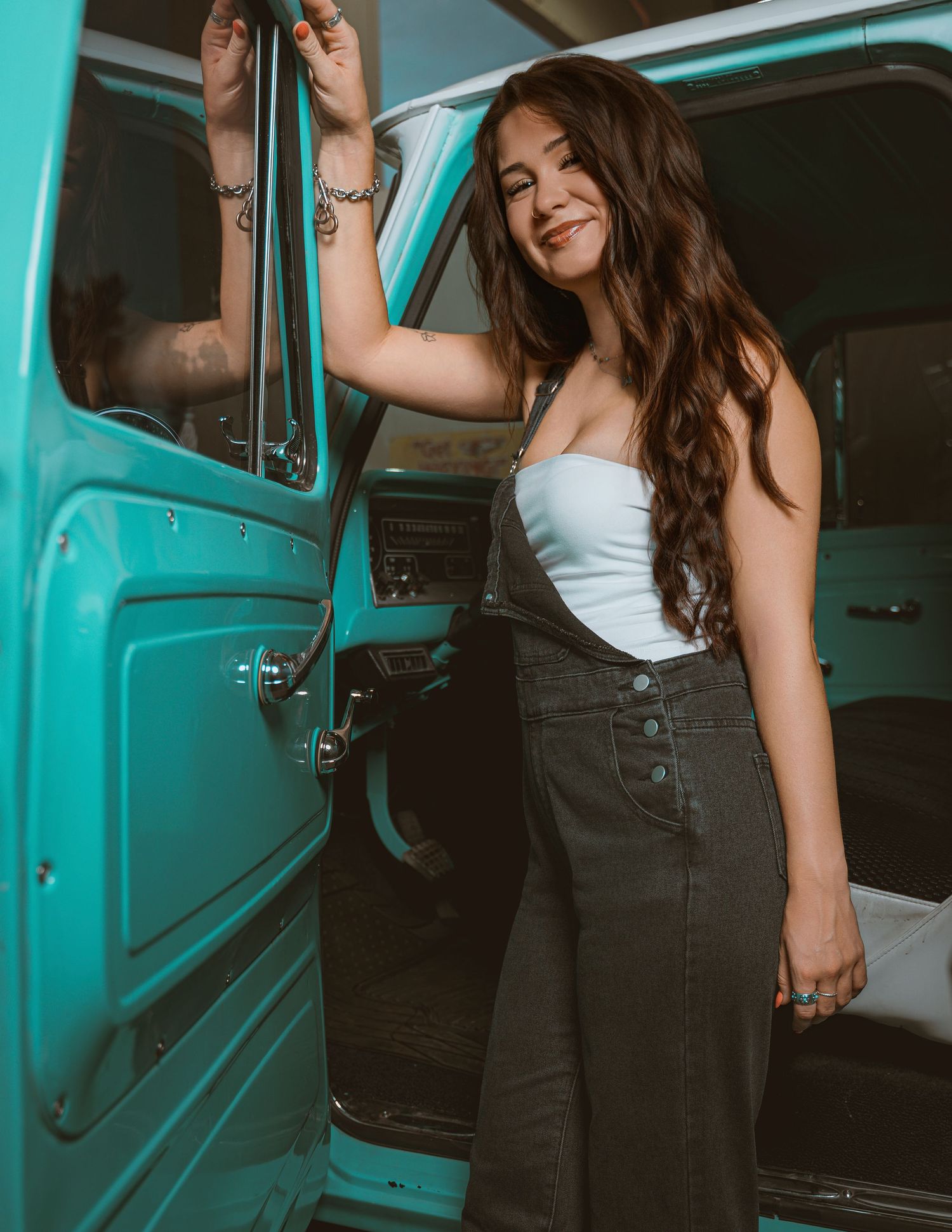 Young woman in white top and black overalls smiling beside a vintage teal truck door.