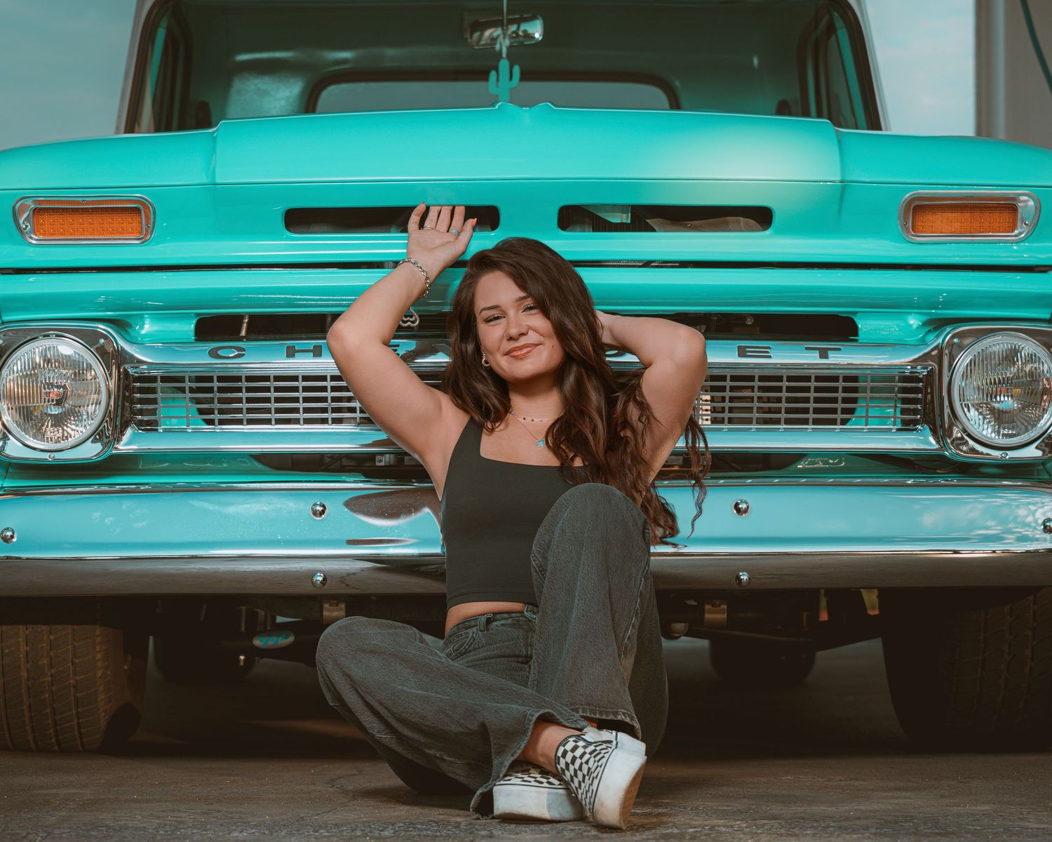 Young woman sitting in front of a vintage turquoise Chevrolet truck wearing a black crop top and gray pants.