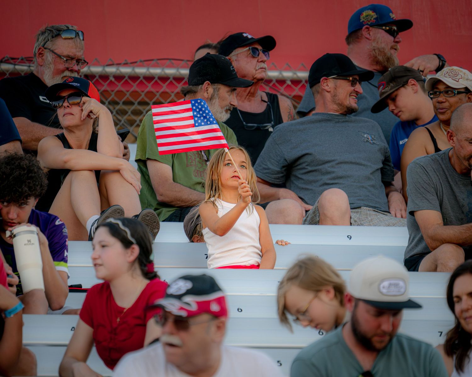 Young girl holds American flag in crowded bleachers at outdoor event, surrounded by spectators on a sunny day.