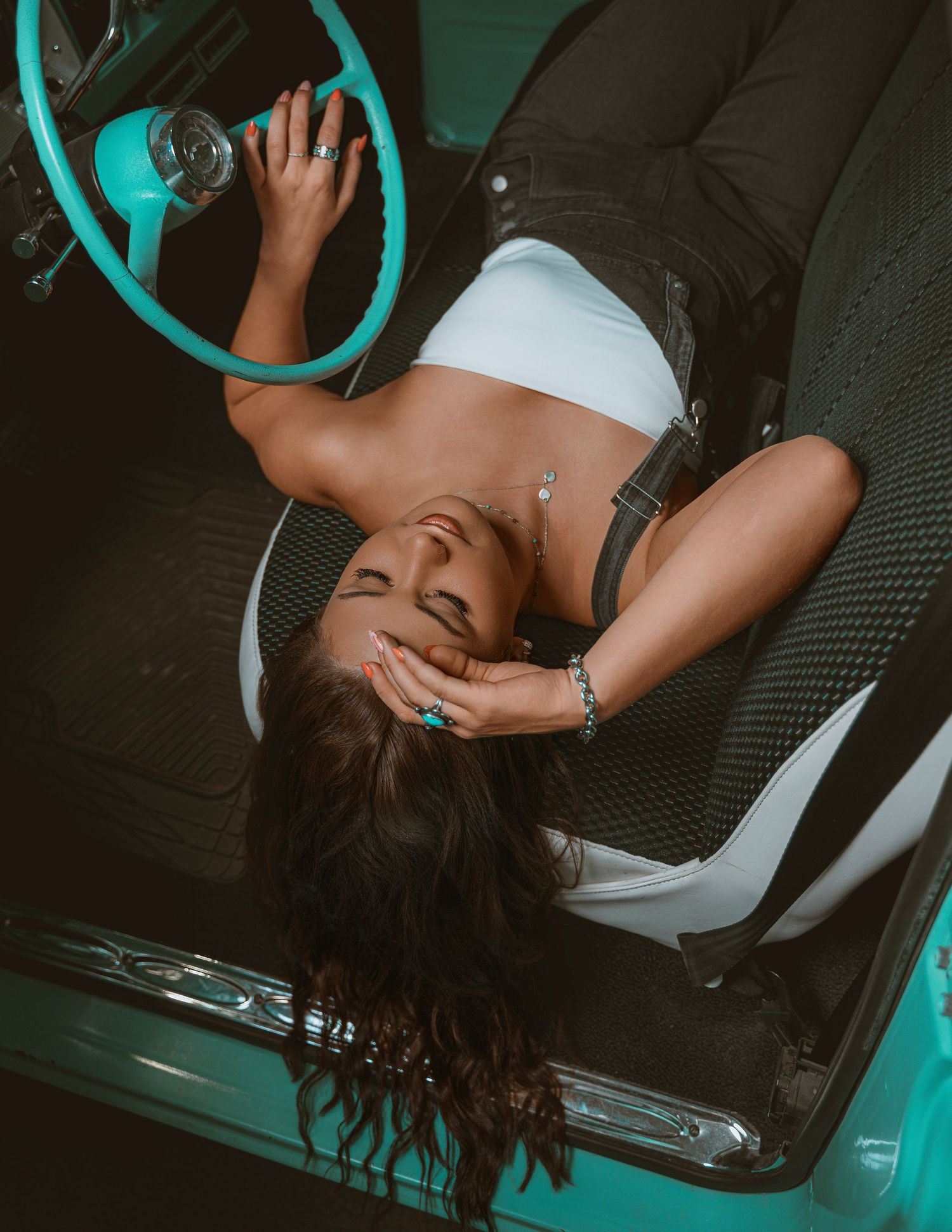 Woman in white crop top reclines in vintage teal car, laughing with hand on face, wearing turquoise jewelry.