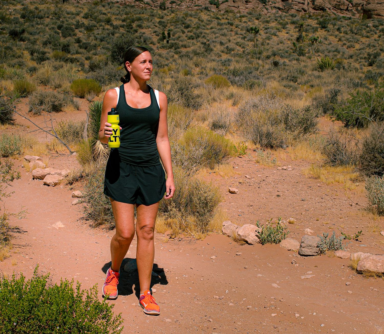 Woman trail running in desert landscape holding yellow LMNT water bottle wearing black athletic outfit and orange shoes.