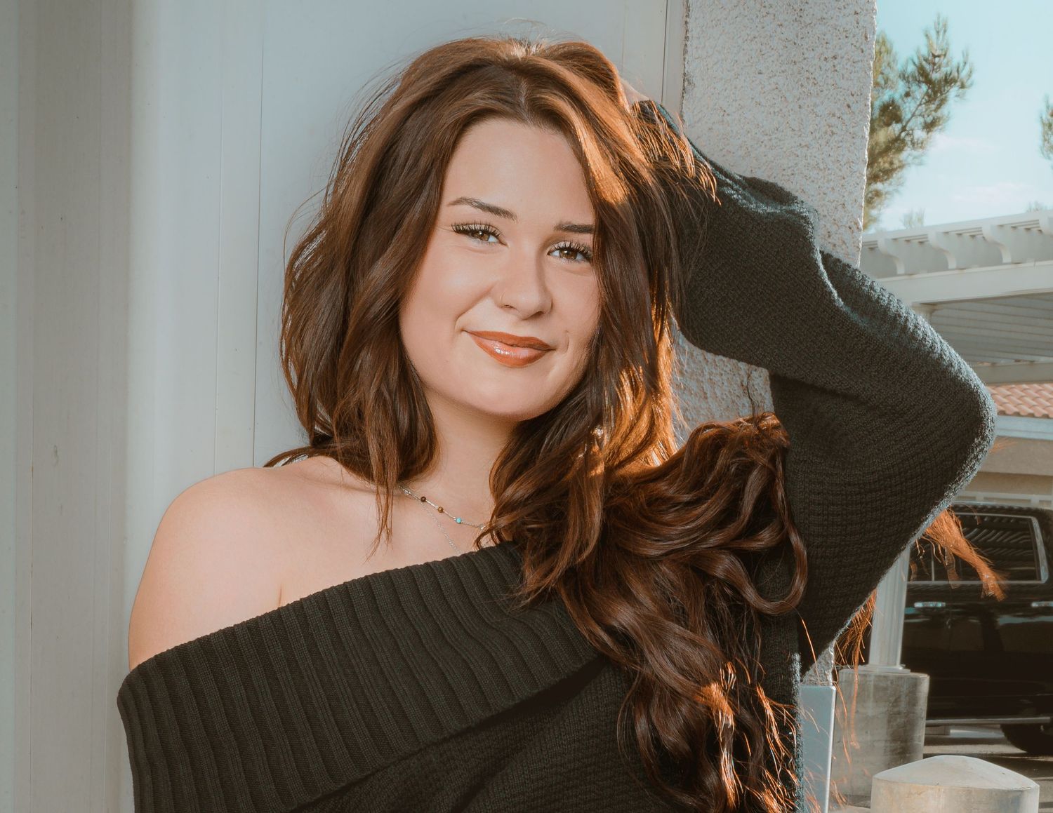 Young woman with long auburn hair wearing an off-shoulder dark green sweater, smiling warmly near a bright window.