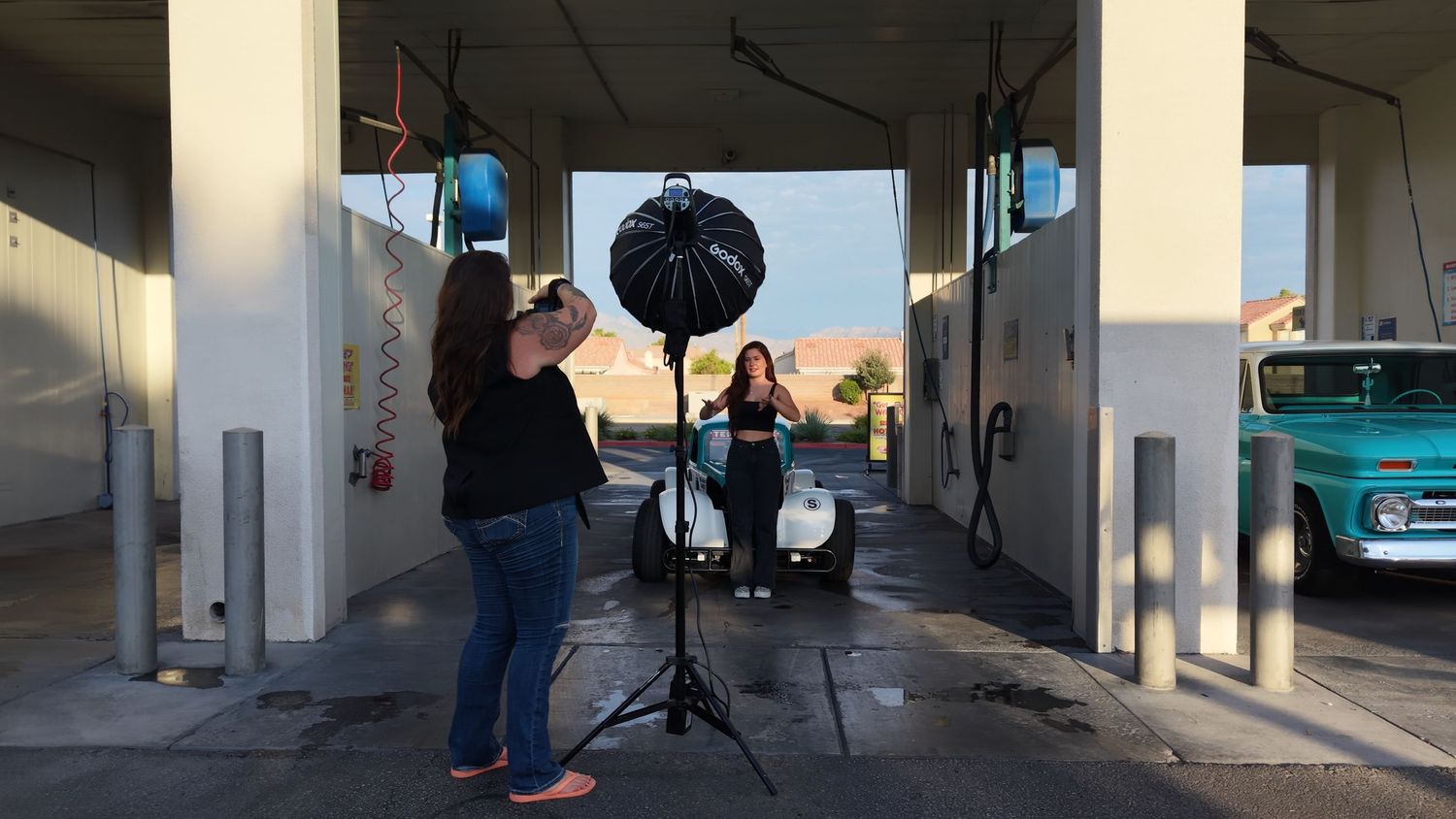 Photographer with lighting equipment shoots a woman posing in an open car wash bay with a vintage truck in background.