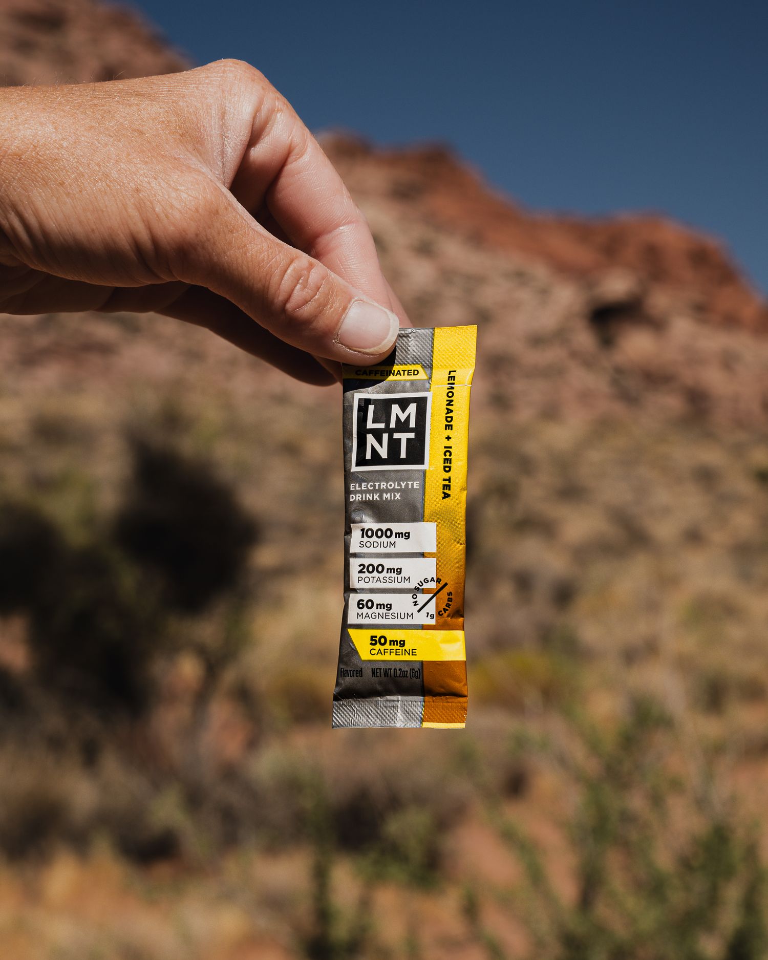Hand holding a yellow LMNT electrolyte drink mix packet with red rock desert landscape in background.