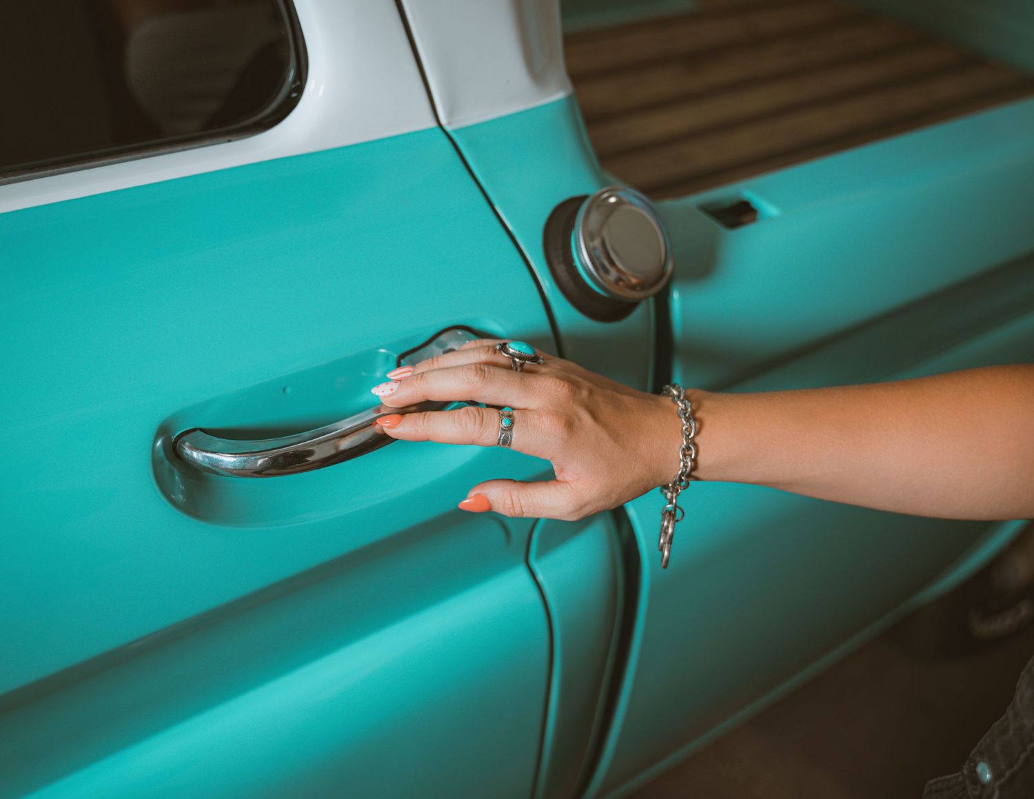 A woman's hand with rings and a bracelet opening a turquoise vintage car door handle.