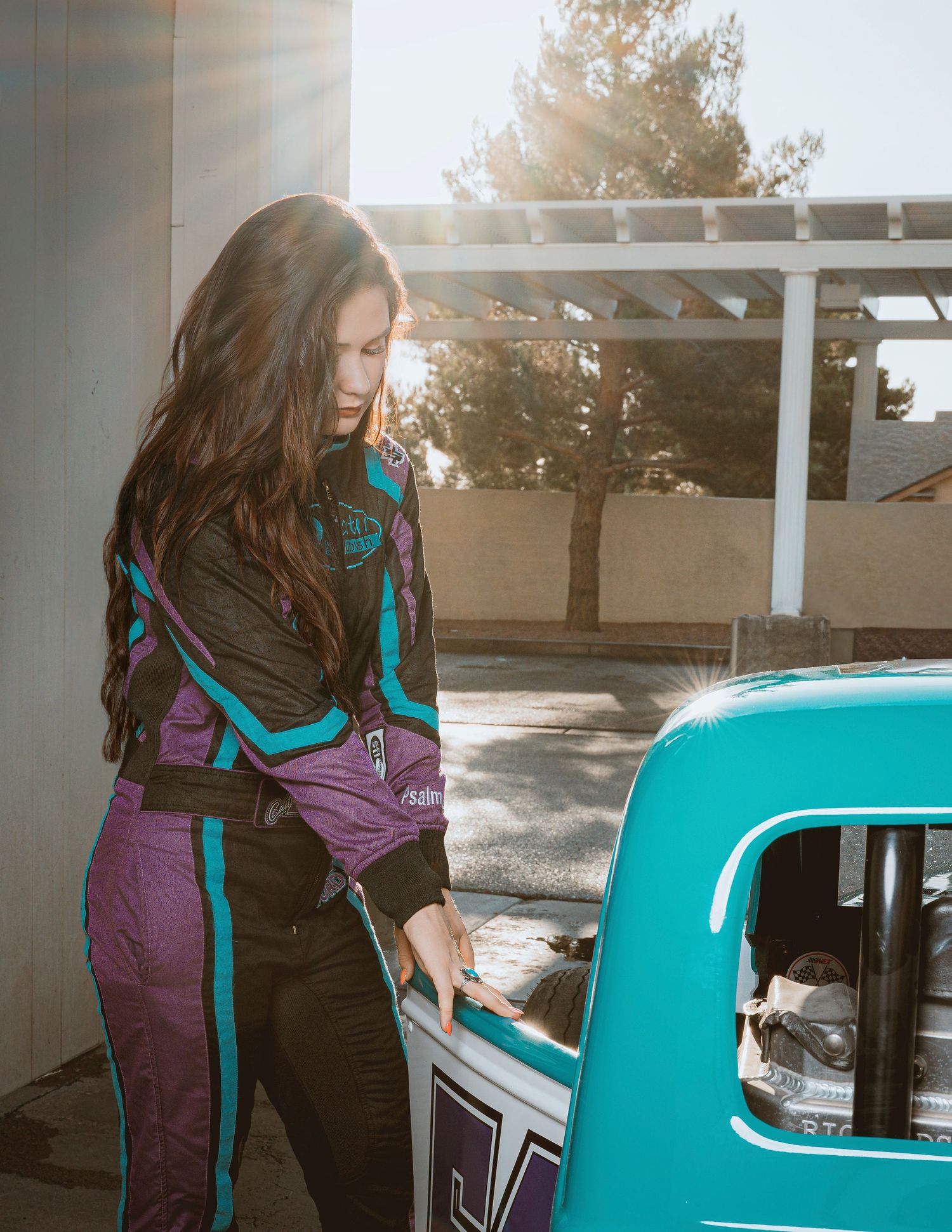Woman in colorful racing suit standing beside a teal race car outdoors.