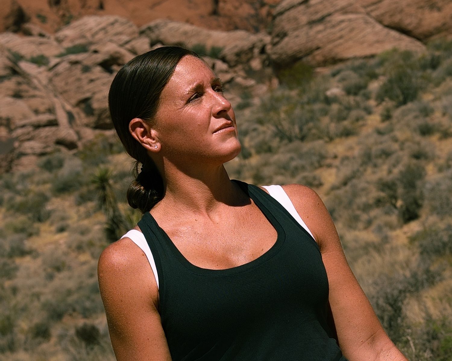 Athletic woman in black tank top looking up while resting outdoors among red rock desert landscape.