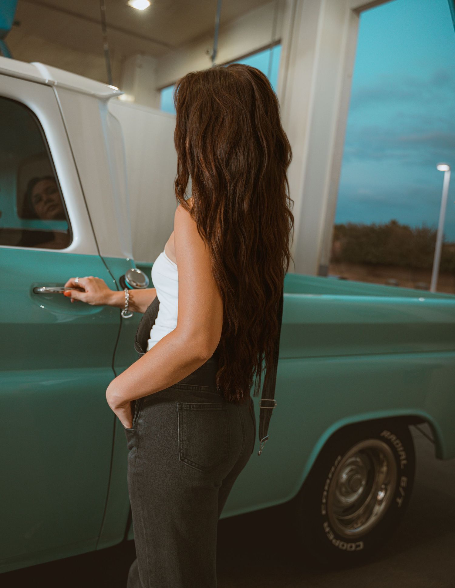 Young woman with long wavy brown hair opening a vintage teal pickup truck door at dusk.