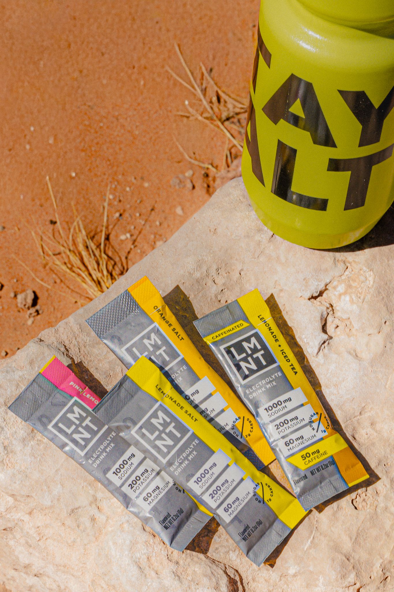 Three LMNT electrolyte drink mix packets displayed on a desert rock beside a yellow STAY SALTY bottle.