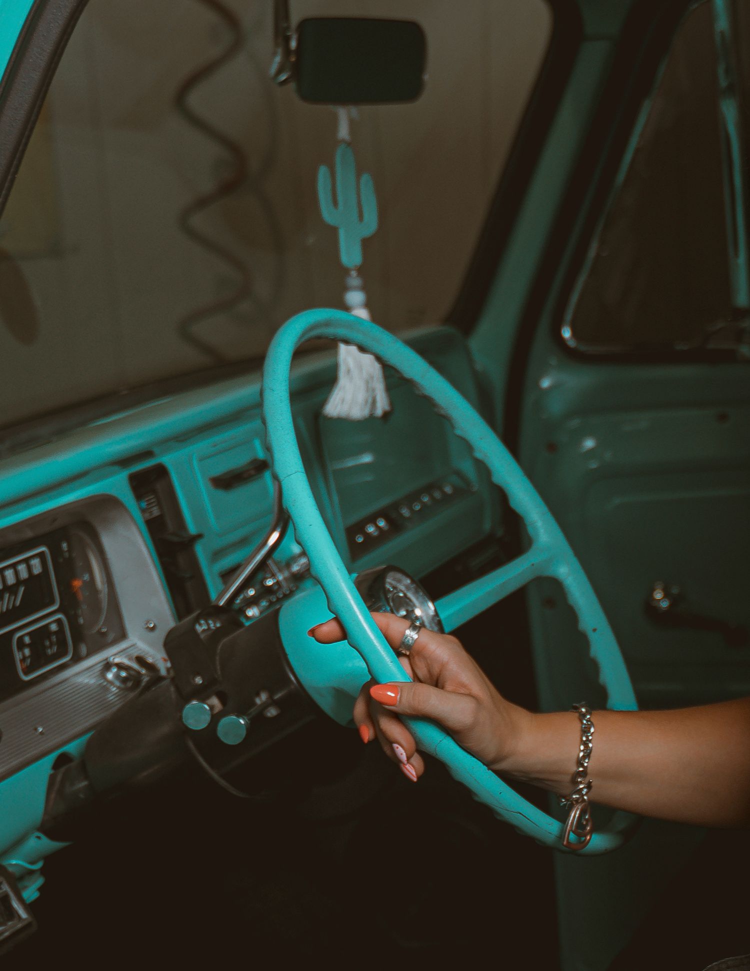 A woman with orange nails and a bracelet grips a turquoise steering wheel inside a vintage teal truck.