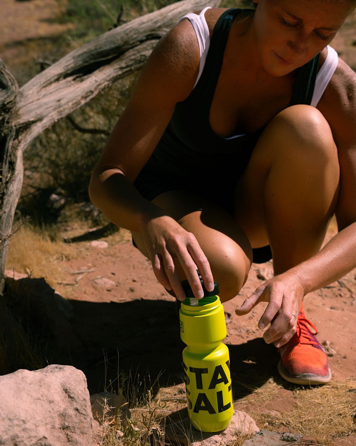 Female trail runner crouching on desert ground preparing yellow LMNT water bottle with orange Nike shoes.
