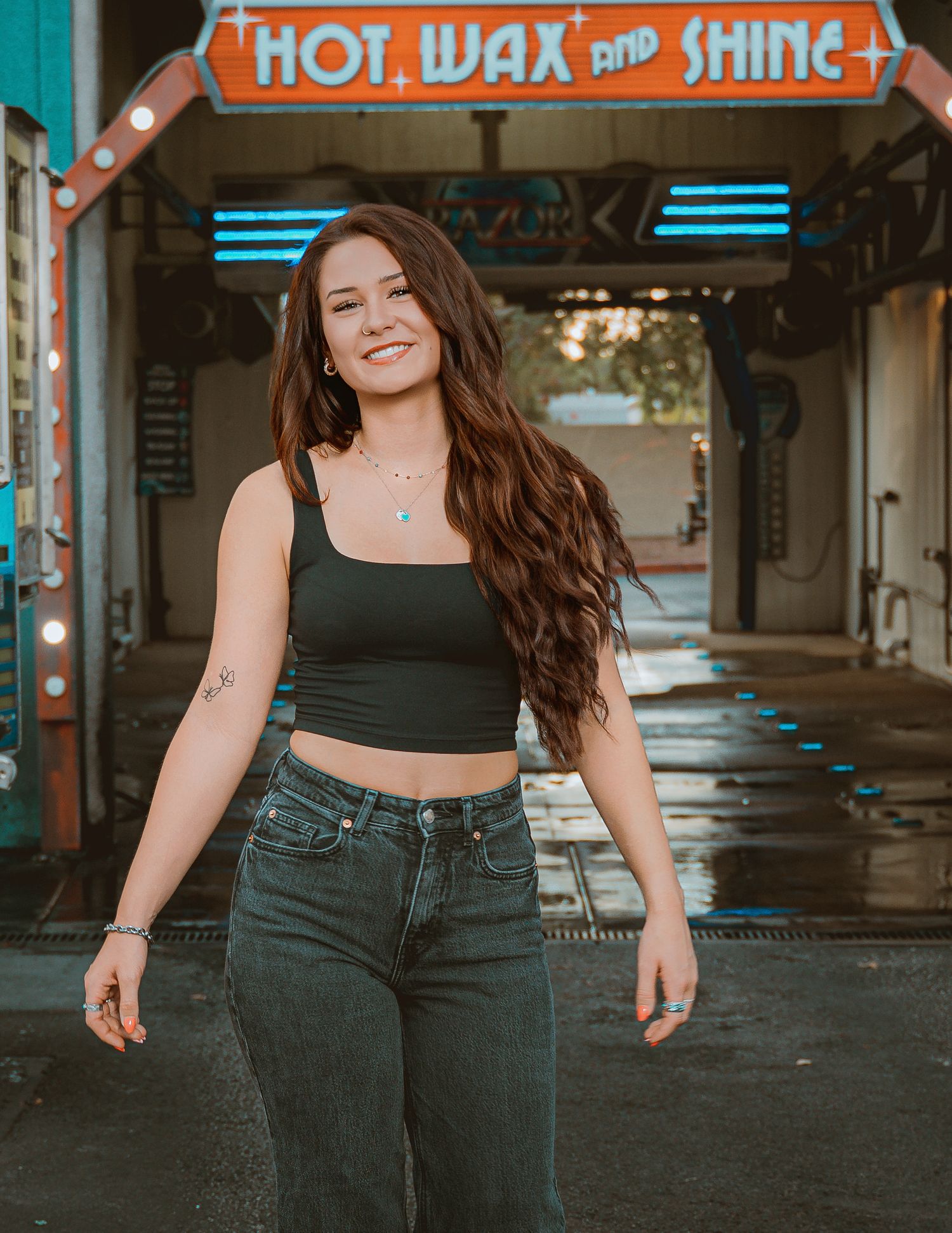 Young woman with long wavy hair in black crop top and jeans smiling outside a Hot Wax and Shine car wash.