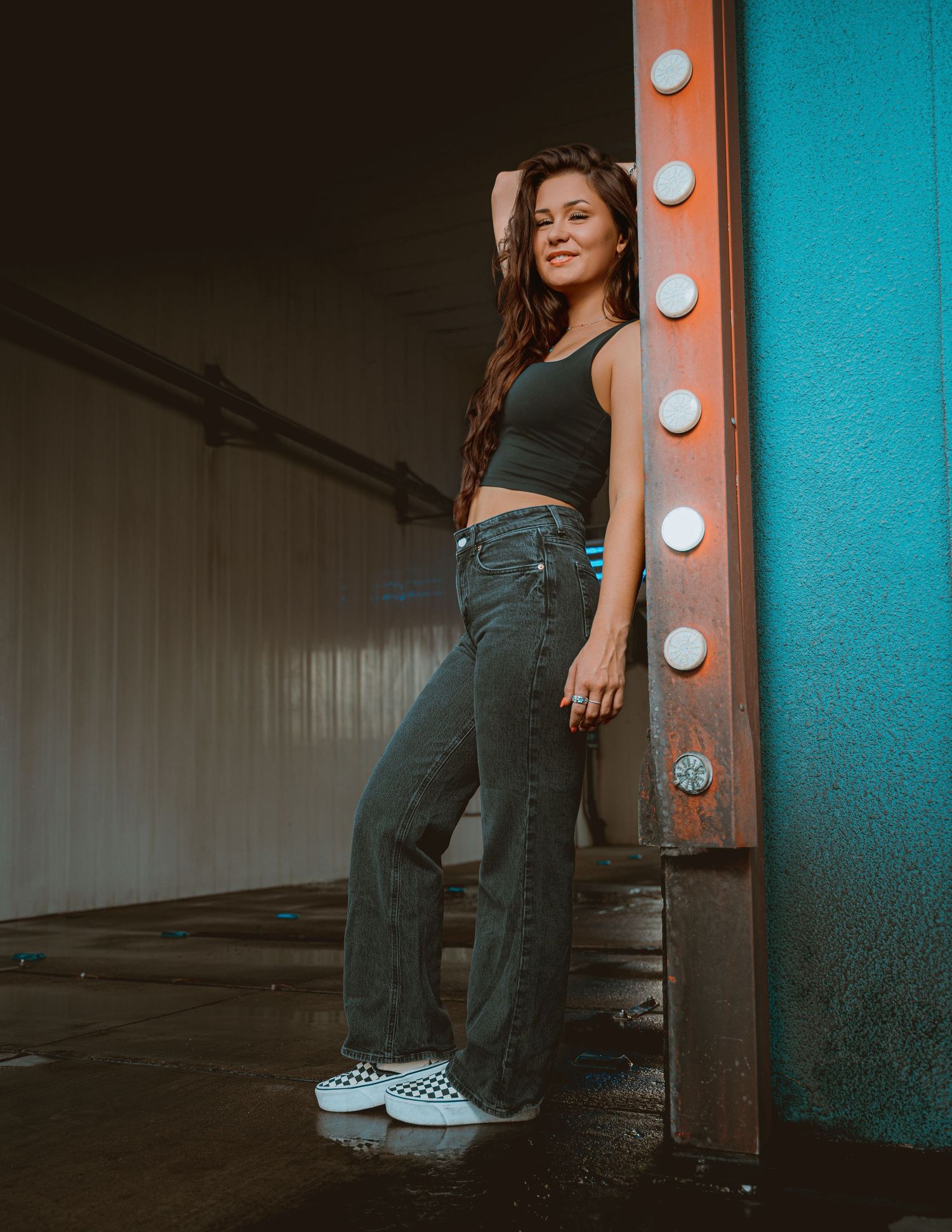 Young woman in crop top, wide-leg jeans, and checkered sneakers poses confidently by illuminated wall in urban setting.