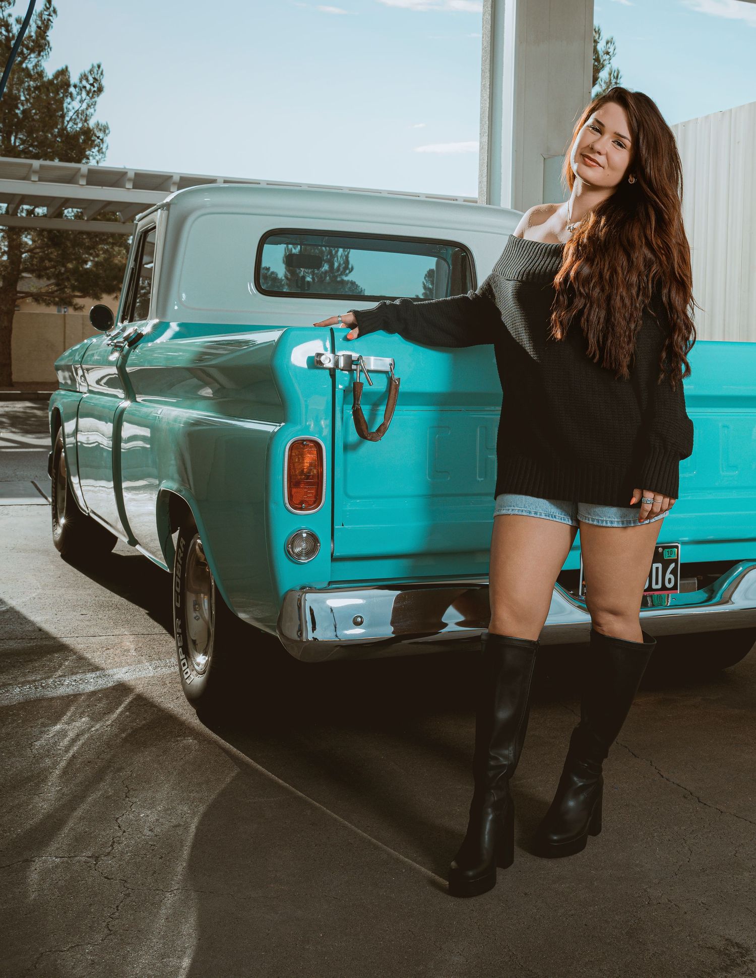 Young woman in black sweater, denim shorts, and knee-high boots posing beside a classic teal vintage pickup truck outdoors.