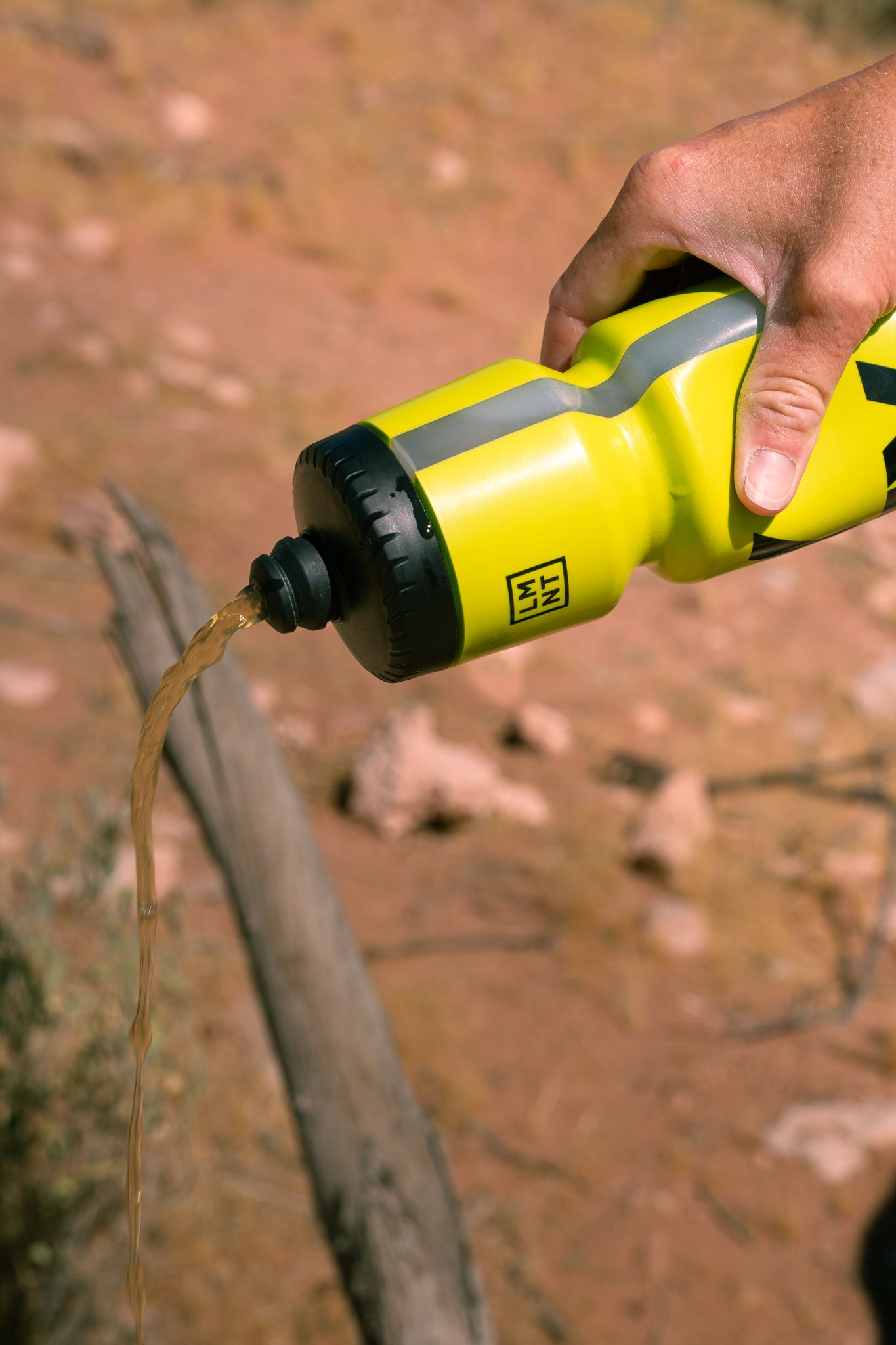 Hand pouring dirty water from yellow LMNT sports bottle onto rocky desert ground outdoors.