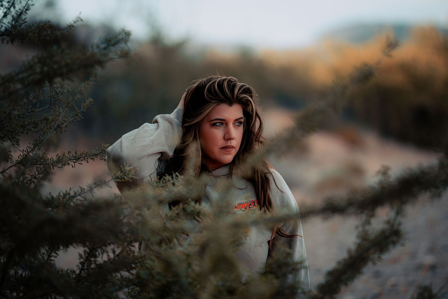 Young woman with dark hair peering through desert shrubs in moody outdoor setting.