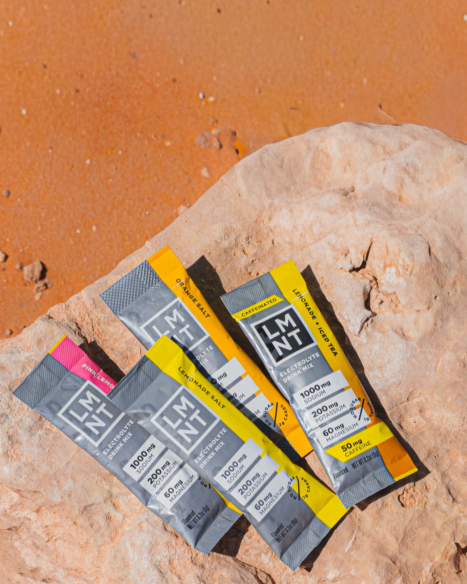 Three LMNT electrolyte packets in citrus and pink lemonade flavors arranged on a sandy desert rock.