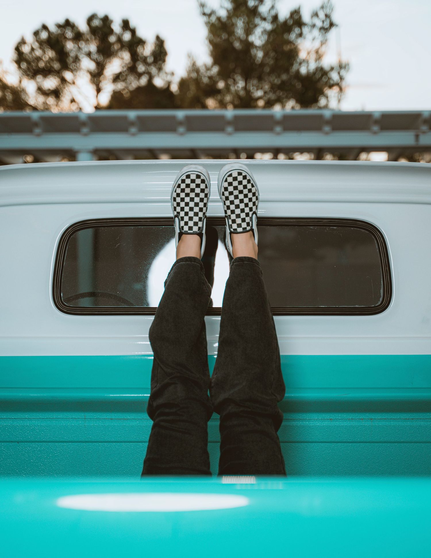 Person wearing checkered Vans slip-on shoes and black jeans, legs propped up on a teal vintage camper van.