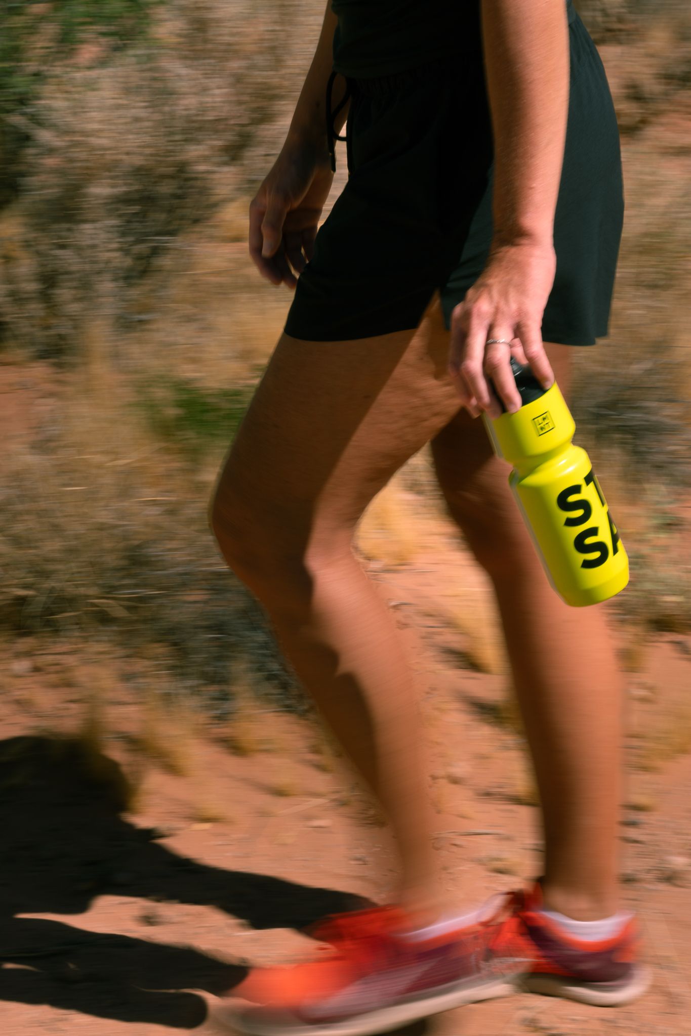 Woman trail running holding a yellow LMNT electrolyte bottle wearing red shoes and black shorts on a desert path.