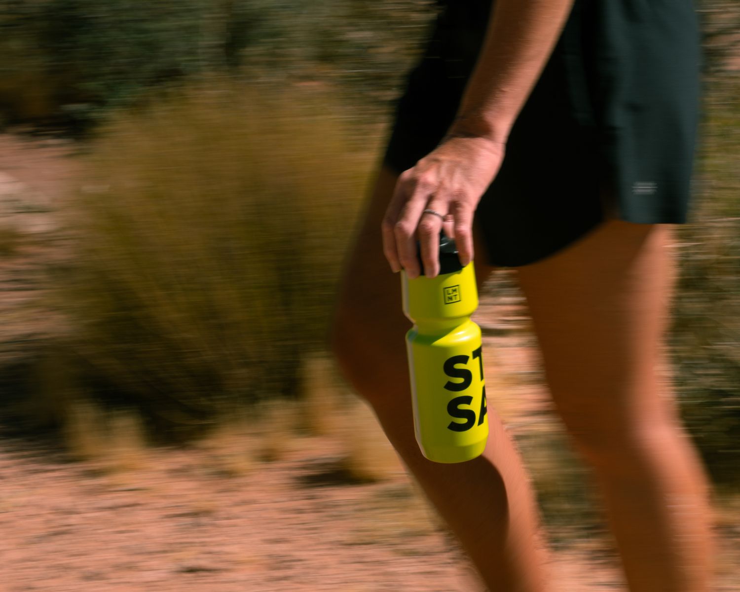Female runner carrying a yellow STAY SALTY LMNT water bottle while running on a desert trail.