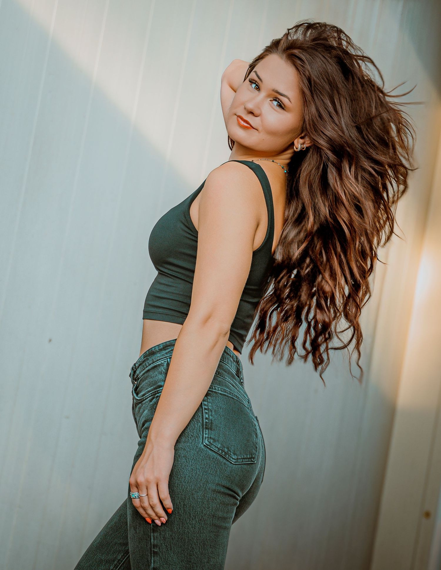 Young woman with long wavy brown hair poses in green crop top and jeans against a light blue wall.