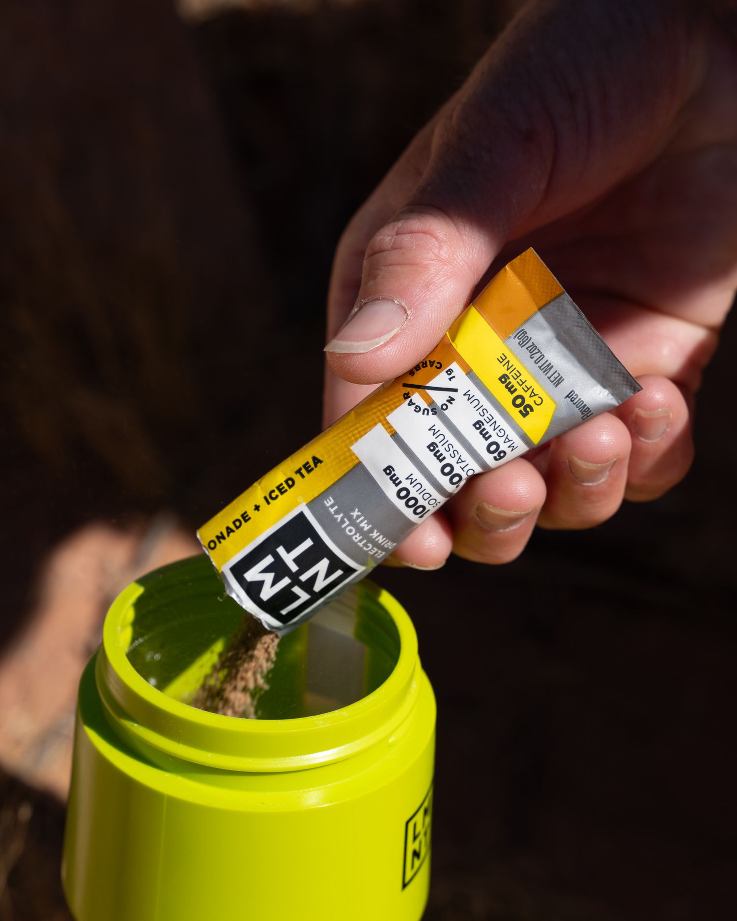 Close-up of hand opening LMNT lemonade iced tea electrolyte packet over yellow LMNT bottle.