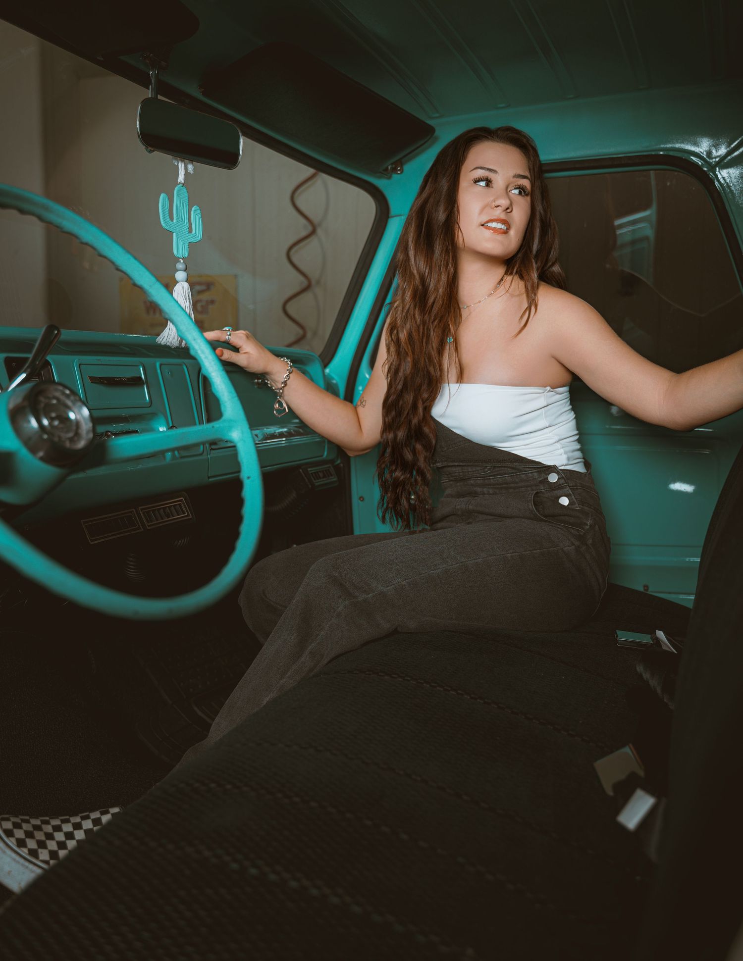 Young woman in white top and black skirt sitting inside a vintage teal truck, posing elegantly.
