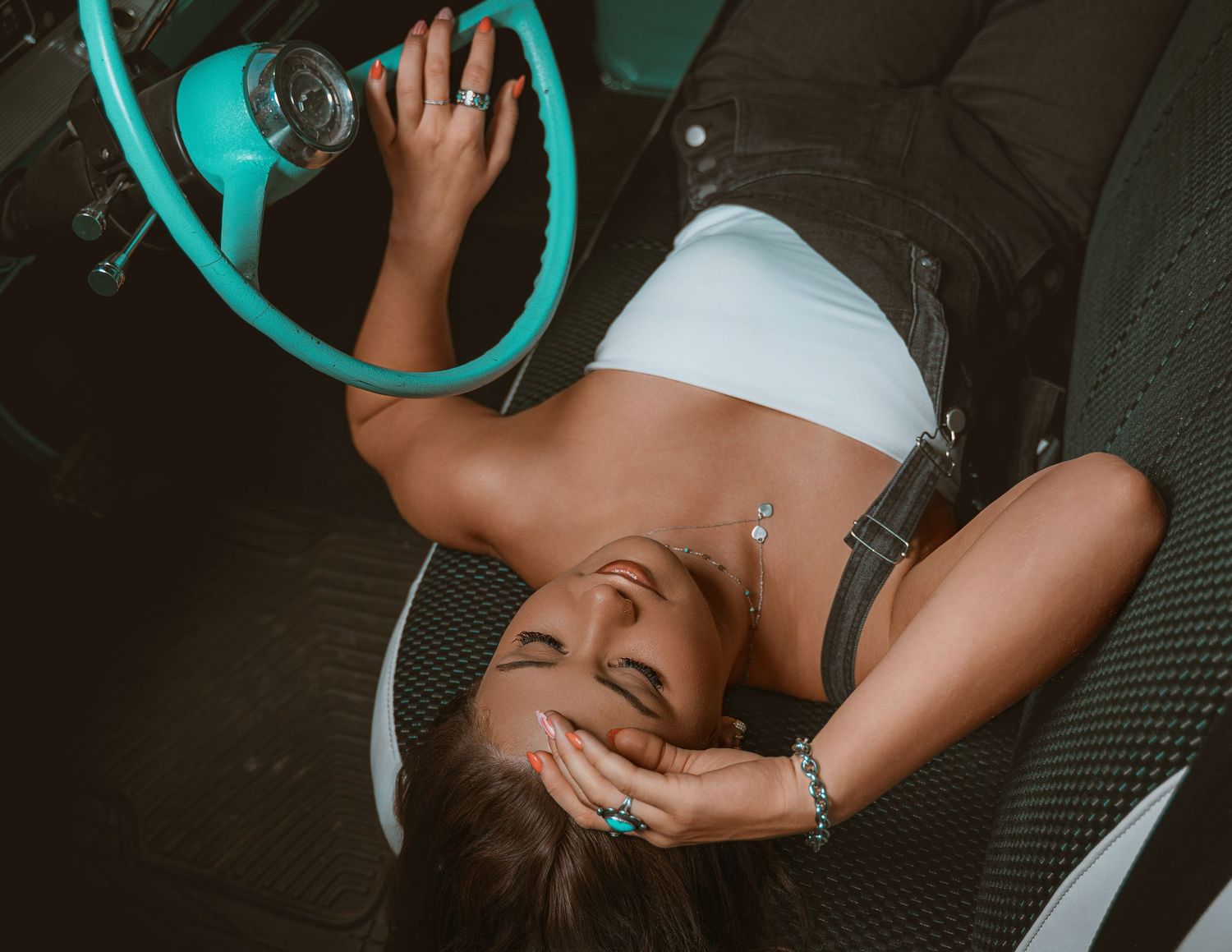 A woman laughing while lying inside a vintage car with a teal steering wheel, wearing a white crop top and overalls.
