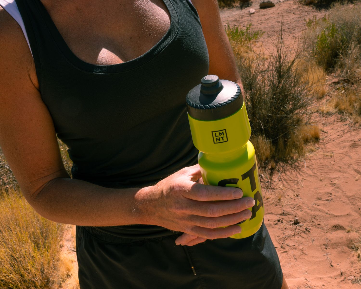 Female athlete in black tank top holding a yellow LMNT branded water bottle on a desert trail.