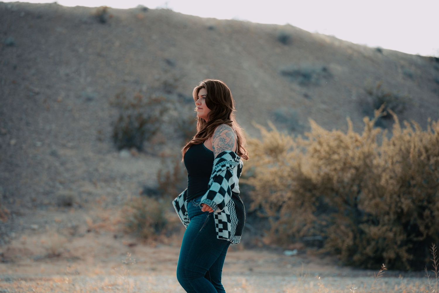 Woman in checkered jacket posing in desert landscape with hills behind her.