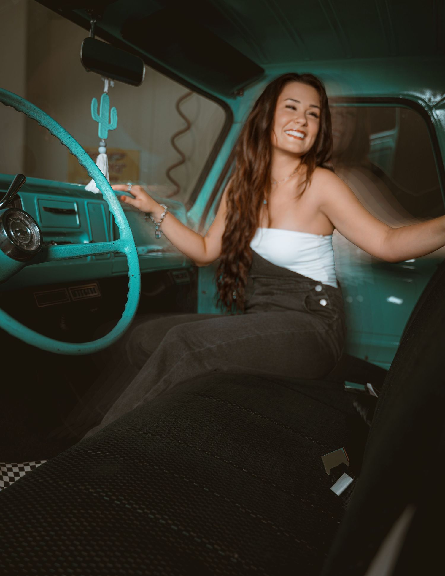 Smiling young woman with long brown hair sitting in a vintage teal truck, wearing a white top and black skirt.