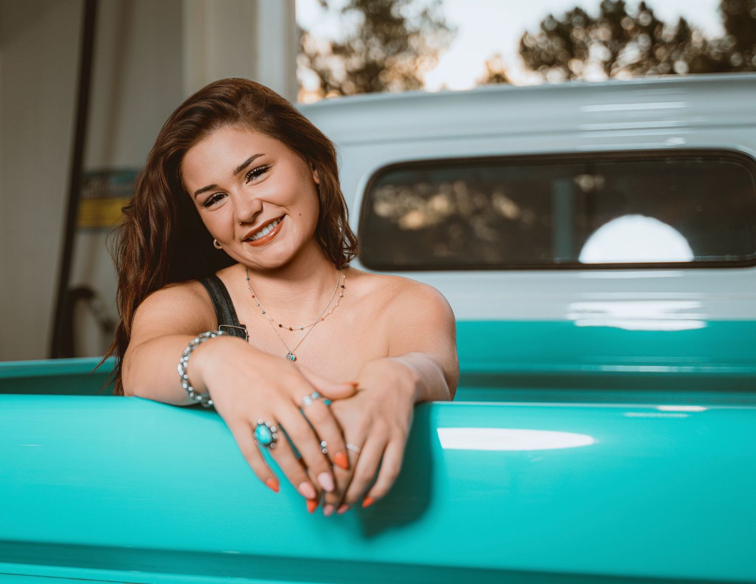 Young woman smiling, leaning on a turquoise vintage truck, wearing jewelry and colorful nails outdoors.