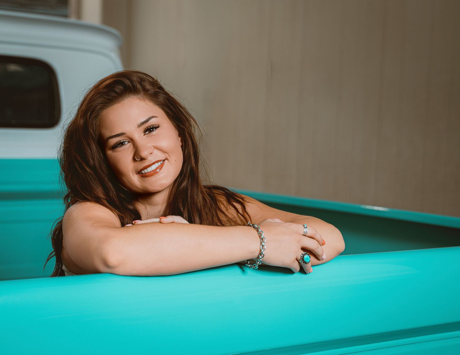 Smiling young woman with long brown hair leaning on a turquoise truck bed, wearing a bracelet.