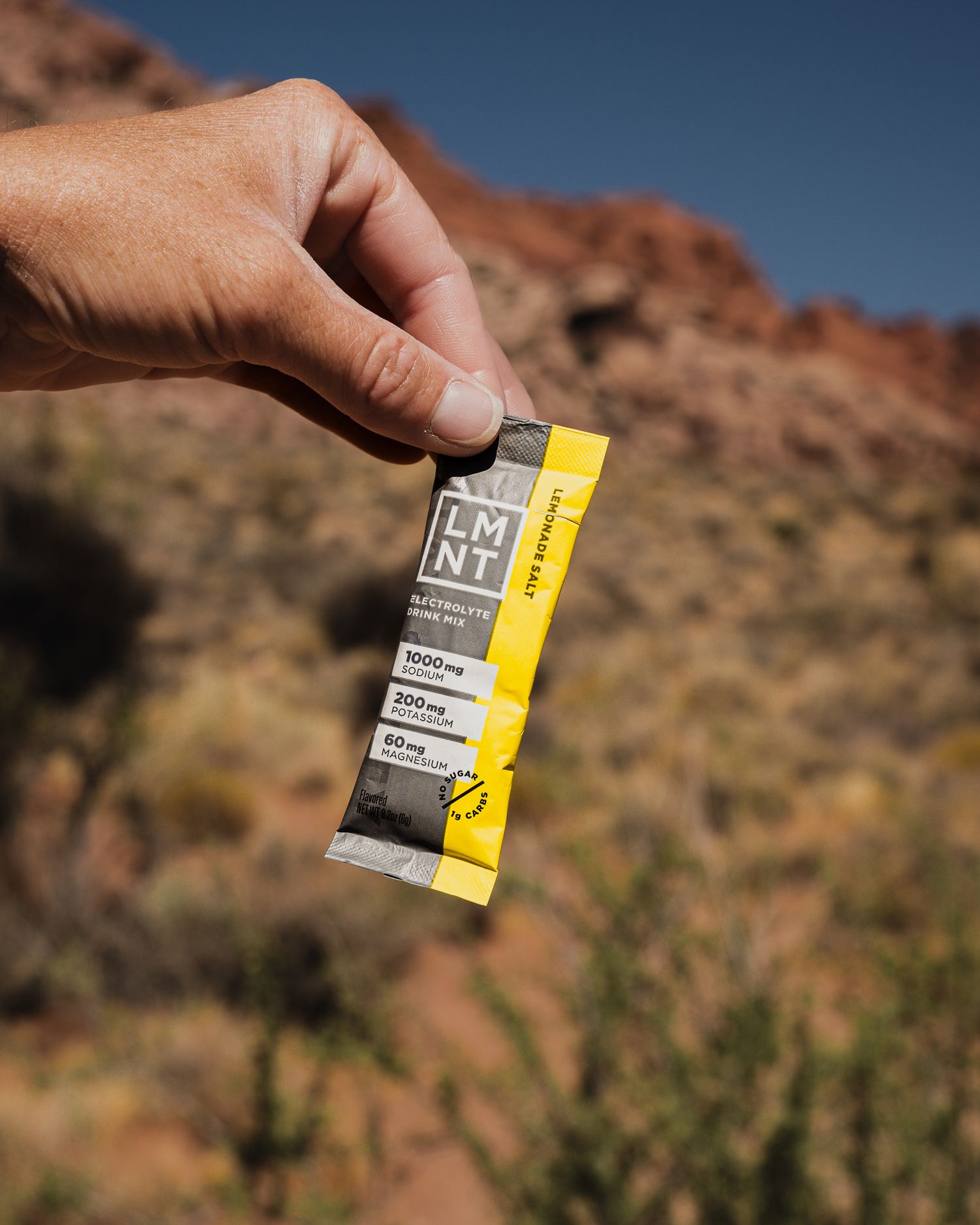 Hand holding LMNT electrolyte drink mix packet outdoors in desert landscape with red rocks.