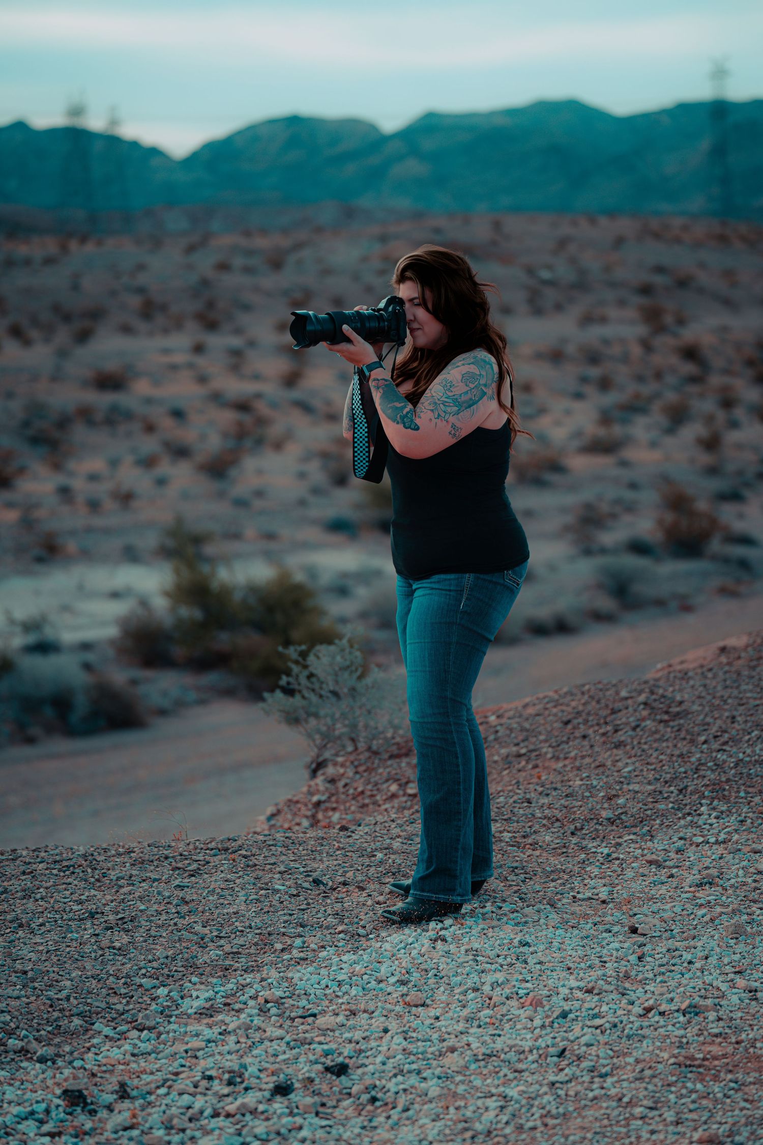 Tattooed woman photographer shooting with DSLR camera in scenic desert landscape at dusk.