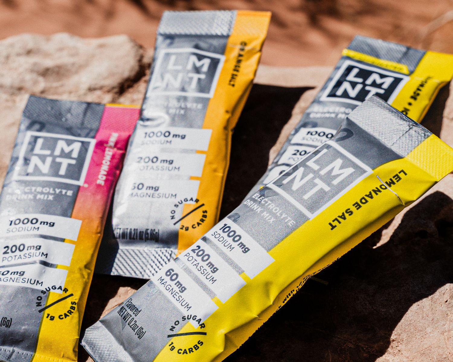 Assorted LMNT electrolyte salt packets in multiple flavors arranged on rocky desert surface.